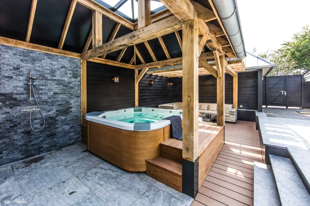Overzicht van de Wellness Garden door ERIC KANT met een luxe hottub, een buitendouche tegen een natuurstenen wand, een houten deck en een sfeervolle loungehoek onder een eikenhouten gebint.