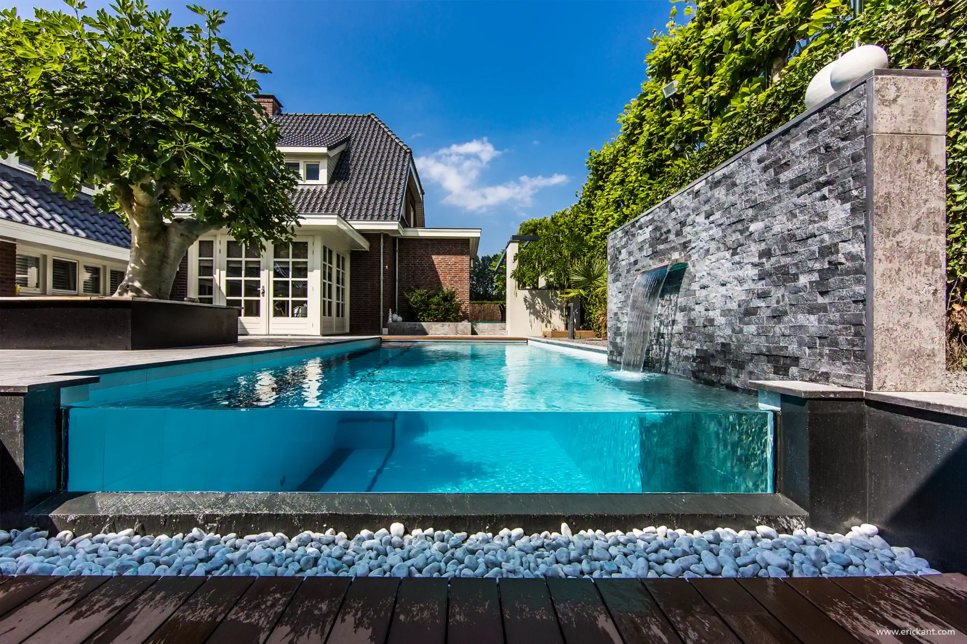 High-end zwembad in de Wellness Garden door ERIC KANT met een transparante glazen zijwand, een waterval uit een natuurstenen muur en een decoratief bed van witte keien.