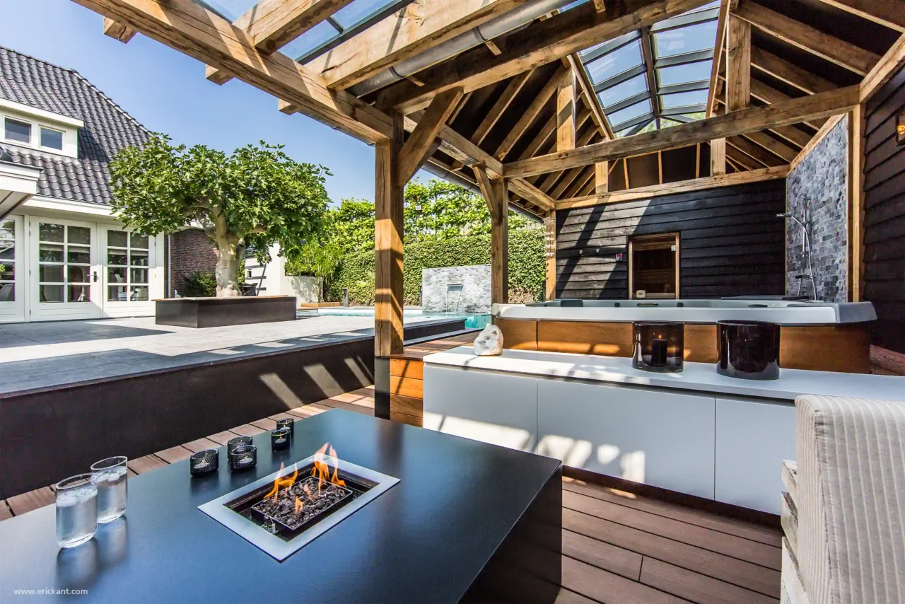 Zicht vanuit de Wellness Garden lounge door ERIC KANT op de achterliggende woning, met de vuurtafel op de voorgrond, de eikenhouten structuur en de moderne tuinarchitectuur.