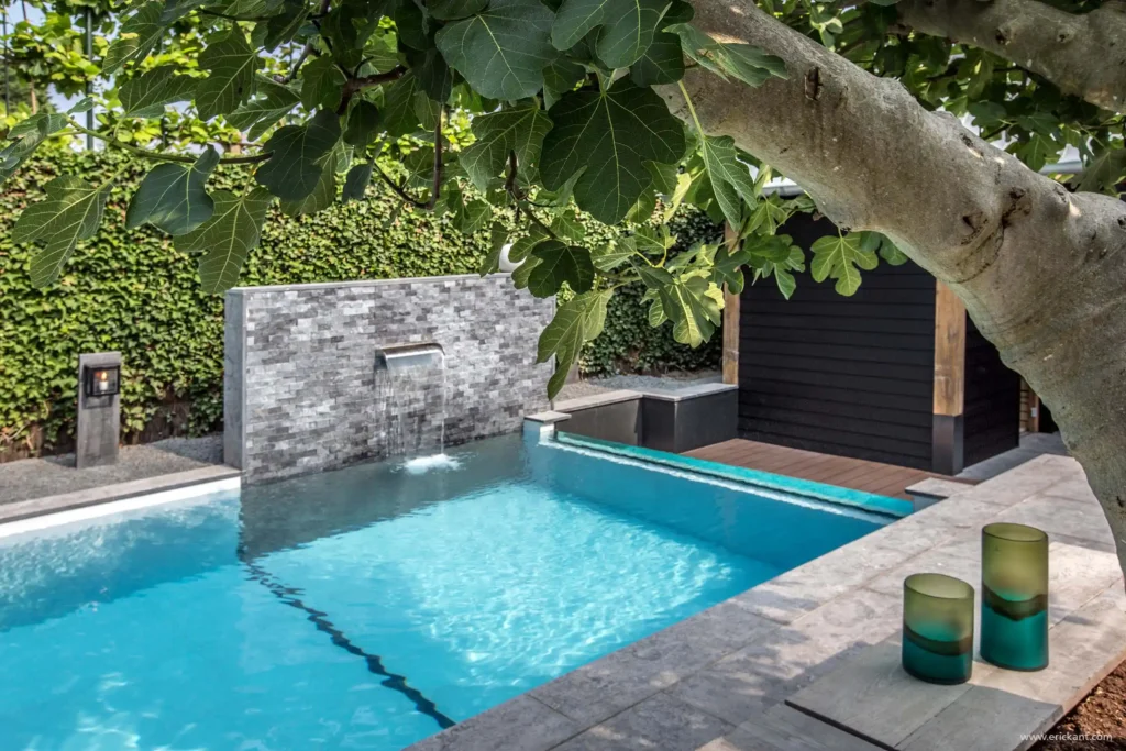 Zicht op het luxe zwembad in de Wellness Garden door ERIC KANT, geframed door de bladeren van een vijgenboom, met de stenen waterval en de glazen zwembadwand op de achtergrond.