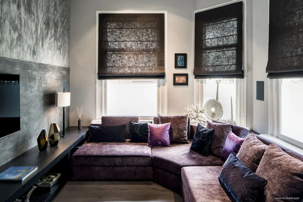 High-end TV-kamer in Riga met paarse loungebank, betonstuc wandafwerking en donkere vouwgordijnen door ERIC KANT.