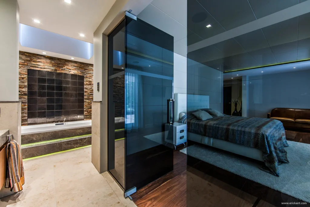 High-end master suite in het Luxury Bungalow project door ERIC KANT met een glazen scheidingswand tussen de slaapkamer en badkamer, een luxe inbouwbad onder een dakraam en rústieke natuurstenen details.