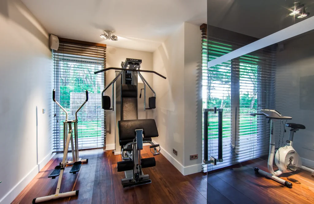 Luxe fitnessruimte in het Luxury Bungalow project door ERIC KANT met een warme houten vloer, moderne fitnessapparatuur, glazen deuren en grote raampartijen met shutters die uitkijken op de tuin.