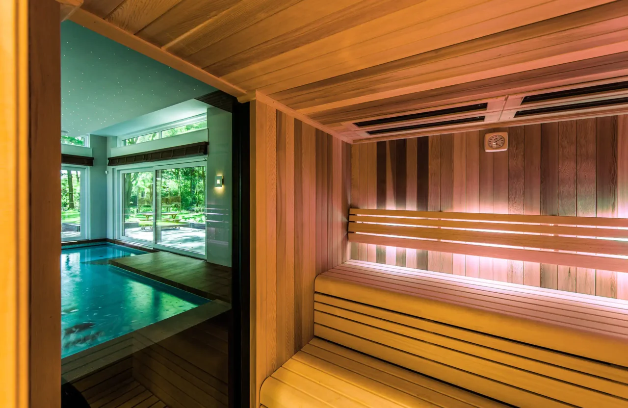 Interieur van een moderne houten sauna in het Luxury Bungalow project door ERIC KANT, met indirecte verlichting en een glazen wand die uitkijkt op het binnenzwembad en de tuin.