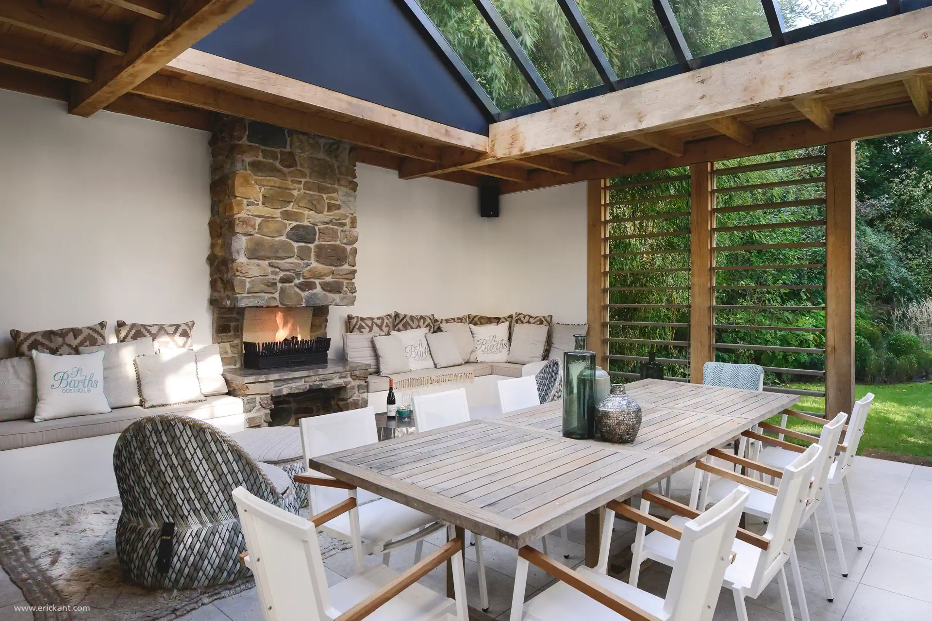 Een luxe overdekt terras ontworpen door ERIC KANT met een robuuste natuurstenen open haard, een grote houten eettafel, witte designstoelen en een glazen daksectie.