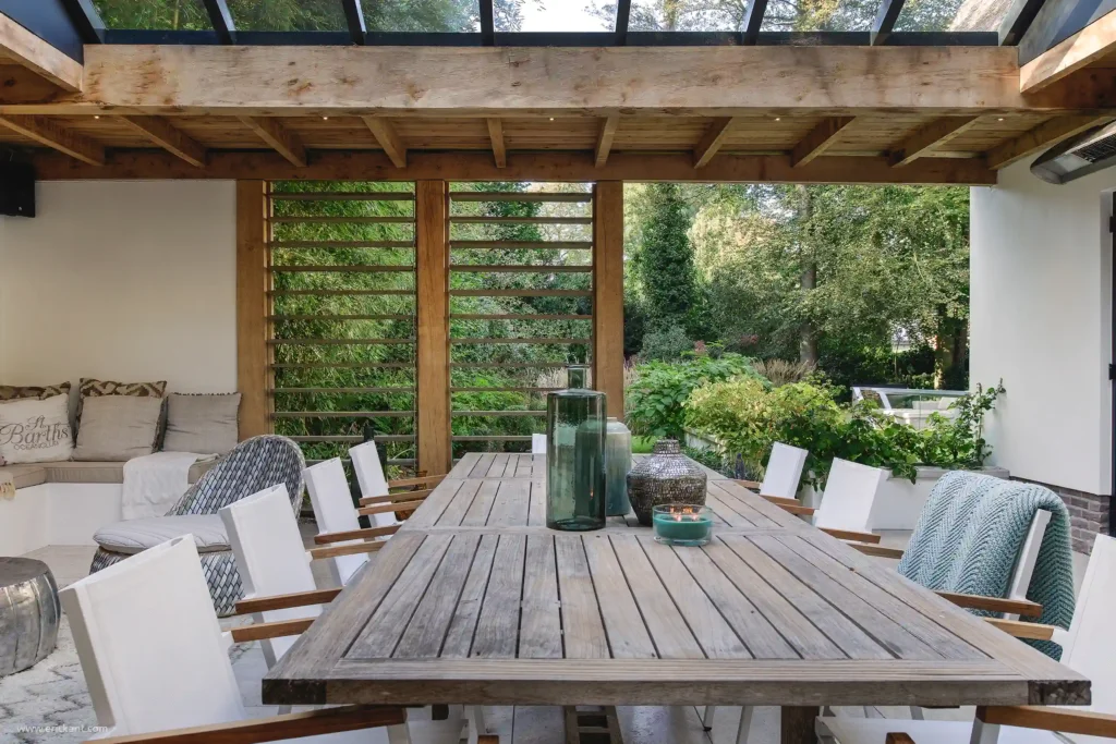 Een luxe overdekt terras ontworpen door ERIC KANT met een rustieke natuurstenen haard, een grote houten eettafel, witte designstoelen en sfeervolle hanglampen onder een donkere houten overkapping.