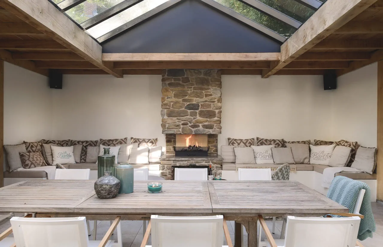 Een luxe overdekte buitenruimte door ERIC KANT met een robuuste natuurstenen open haard, een glazen daksectie in een houten constructie en een eethoek met loungebanken.