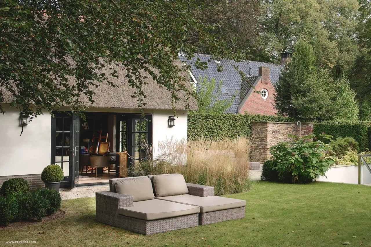 Een luxe dubbel loungebed op een groen gazon bij een rietgedekte villa in Laren, ontworpen door ERIC KANT, met uitzicht op een openstaand atelier en een natuurstenen wellness-muur.
