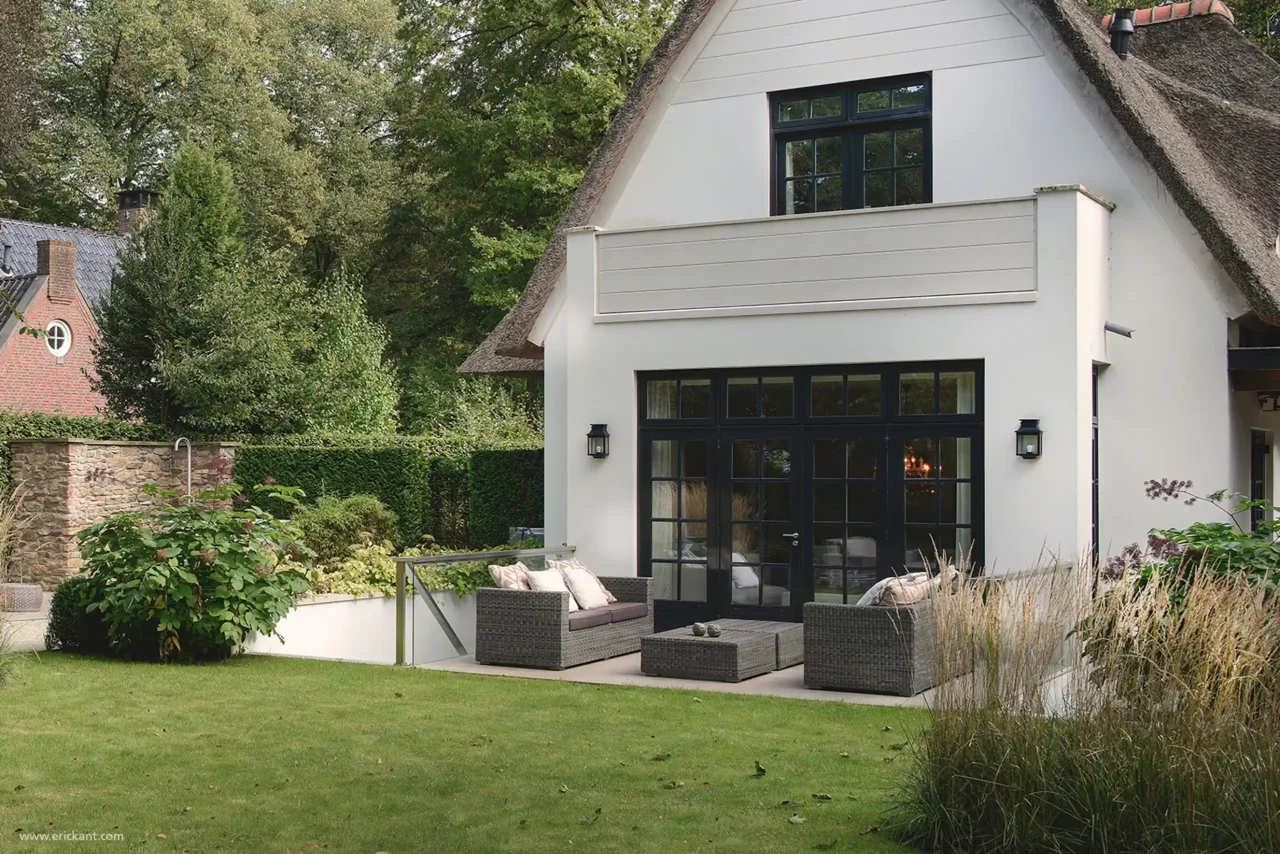 Achteraanzicht van een luxe villa in Laren door ERIC KANT met een rietgedekt dak, witte gevel, zwarte openslaande deuren en een terras met een grijze loungehoek in een groene tuin.