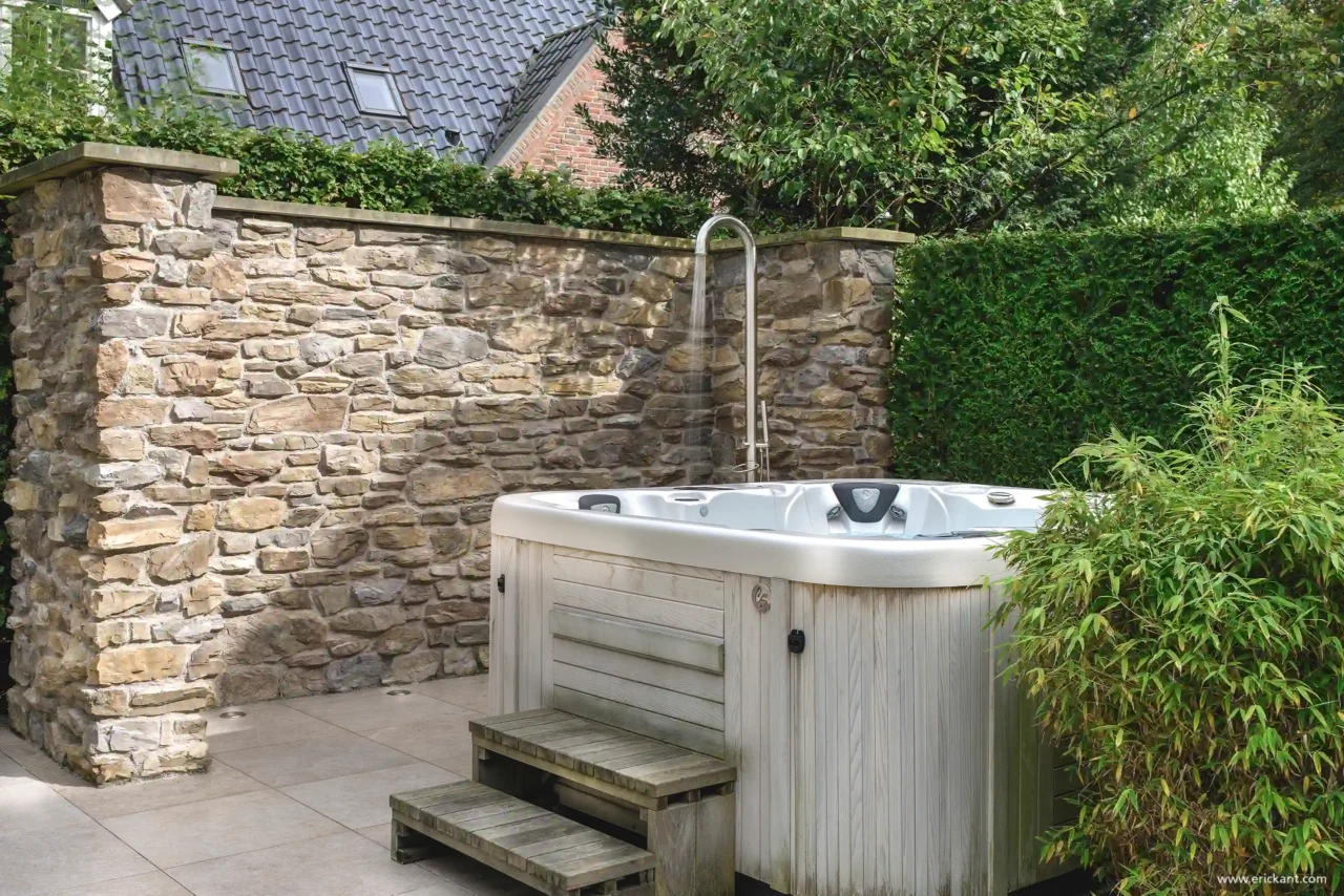 Een luxe jacuzzi met houten ombouw geplaatst voor een robuuste natuurstenen muur, inclusief een moderne rvs buitendouche in de tuin van een villa in Laren door ERIC KANT.