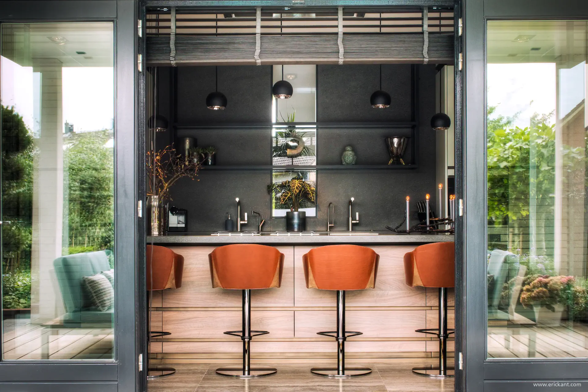 High-end bar in de Mediterranean Holland villa door ERIC KANT, gezien door openstaande tuindeuren, met een licht houten onderstel, cognac lederen krukken, een donkere achterwand en design hanglampen.