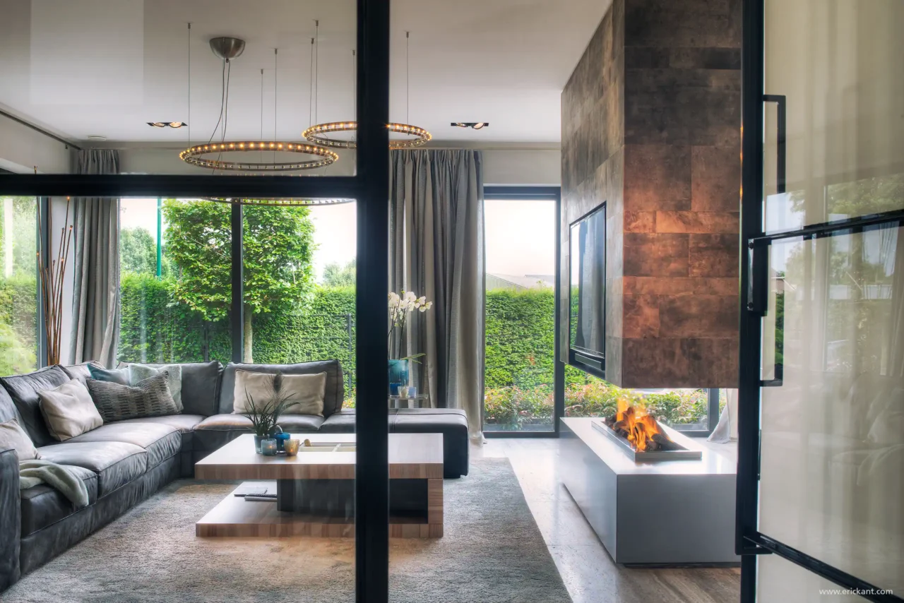 High-end living van de Mediterranean Holland villa door ERIC KANT met een moderne hoekbank, een doorkijkhaard bekleed met bronzen panelen, design ringverlichting en zicht door zwarte stalen deuren.