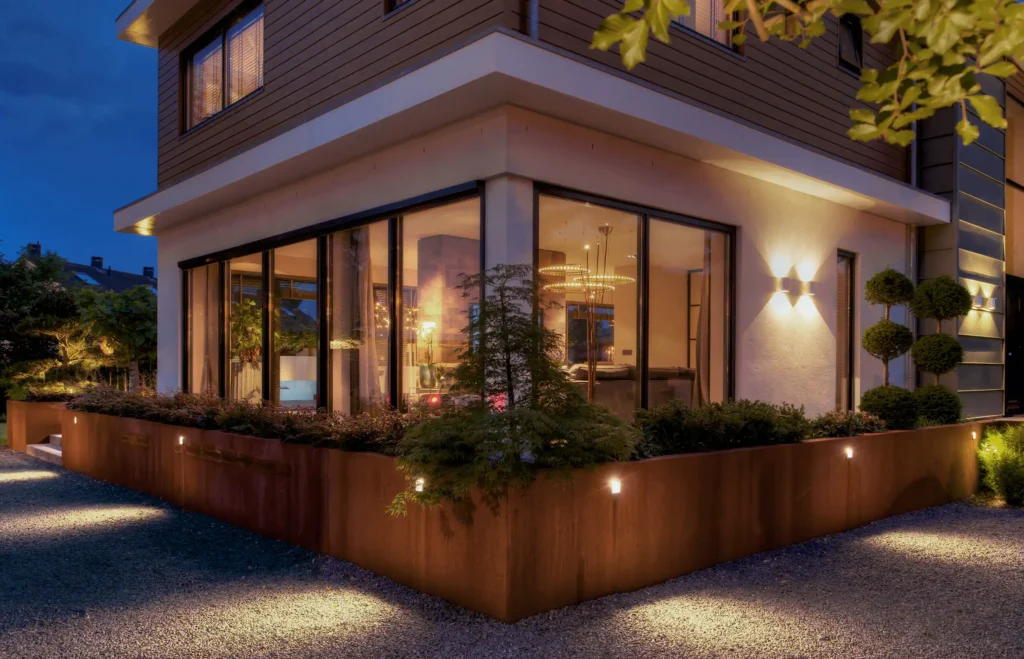 Avondopname van de Mediterranean Holland villa door ERIC KANT met cortenstaal plantenbakken, een grindpad en grote glazen hoekpartijen die zicht bieden op de verlichte living en designringlampen.