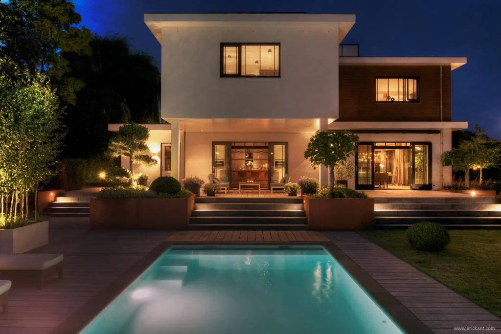 Achteraanzicht van de Mediterranean Holland villa door ERIC KANT bij avond, met een verlicht buitenzwembad, houten decking, cortenstaal plantenbakken en zicht op de bar en dining door de glazen puien.