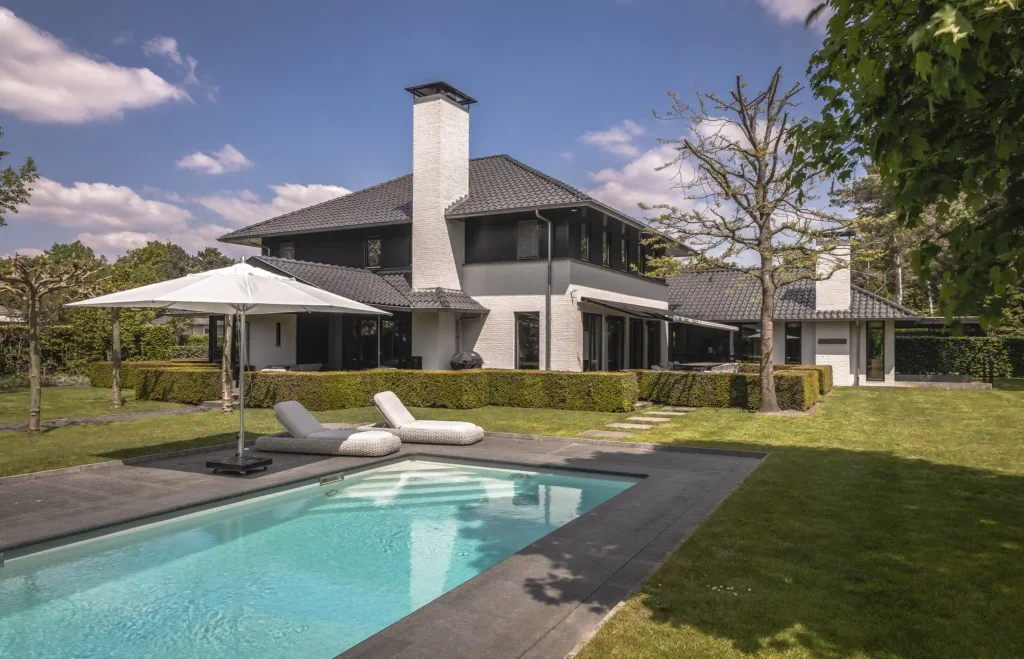 Een luxe zwembad met antracietkleurige rand, witte ligbedden en parasol in de tuin van de Renovation Villa door ERIC KANT, met uitzicht op de wit-zwarte gevel.