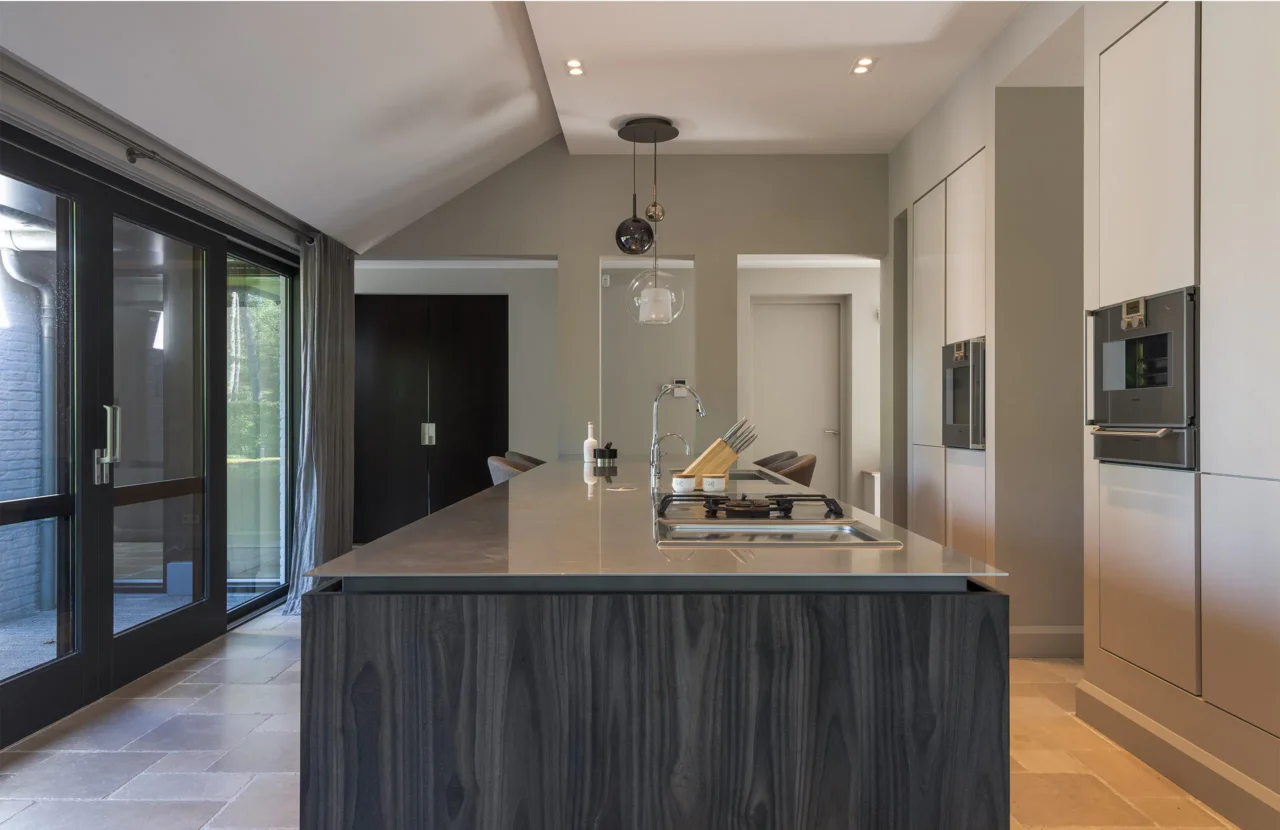 Een luxe keukenontwerp door ERIC KANT met een groot spoeleiland van donker hout, een dun stenen werkblad, geïntegreerde ovens en grote zwarte schuifdeuren naar het terras.