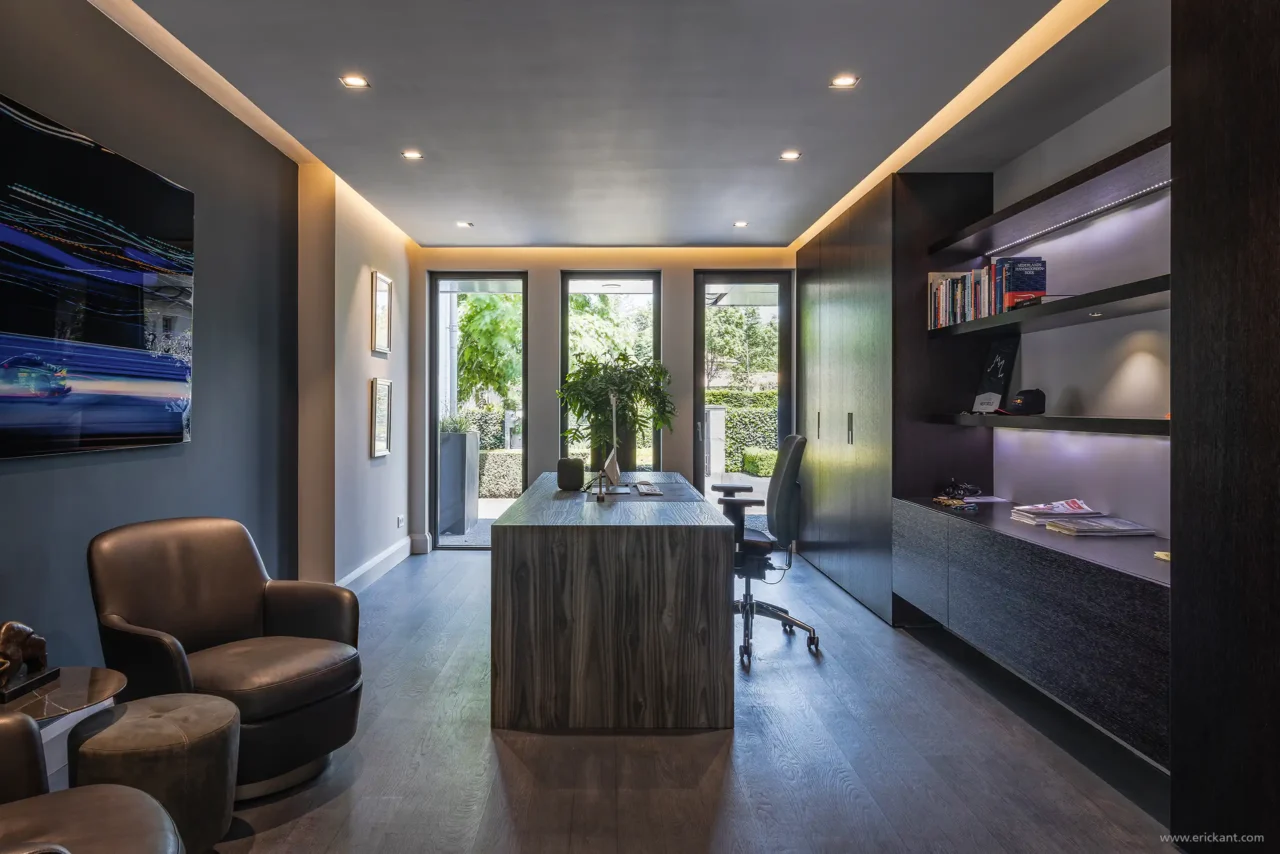 Luxe werkkamer door ERIC KANT met een robuust houten bureau, maatwerk kastenwand met geïntegreerde verlichting, donkere wanden en uitzicht op de tuin.