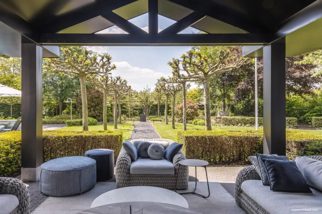 Zicht vanaf een overdekt loungeterras door ERIC KANT met luxe vlechtwerk meubels en blauwe kussens, uitkijkend op een symmetrische tuin met een centraal pad en geknotte bomen.