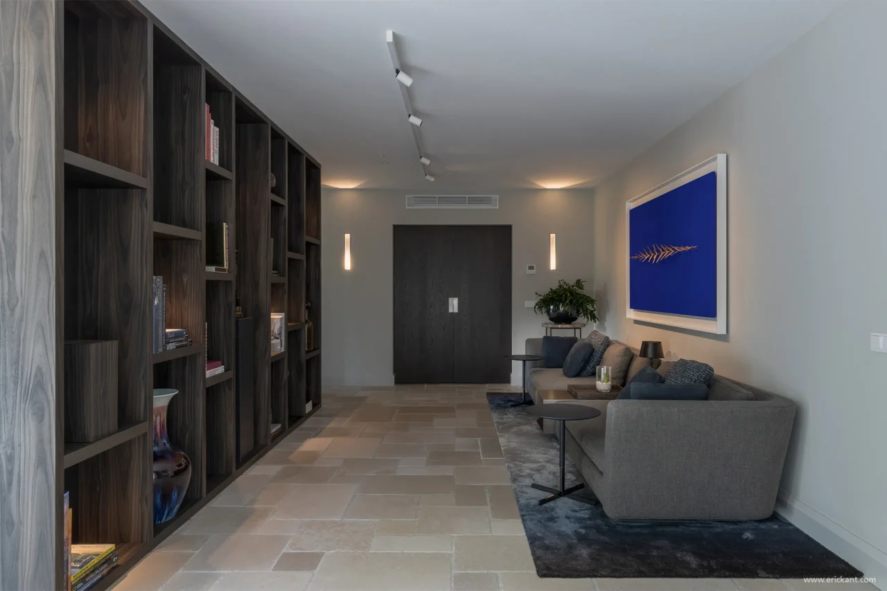 Interieur van een sfeervolle tv-kamer door ERIC KANT met een kamerhoge houten vakkenkast, een comfortabele grijze bank, een blauw kunstwerk en een natuurstenen vloer.