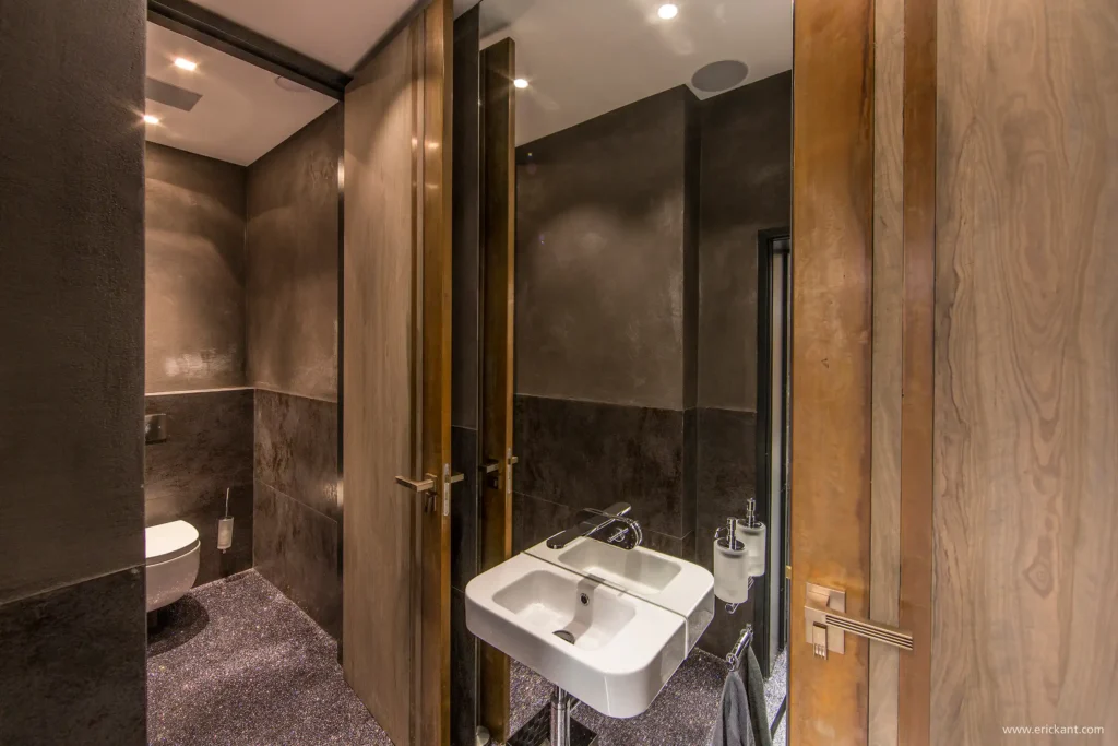 Interieur van de sanitairruimte in de Bowers & Wilkins Concept Store Antwerpen door ERIC KANT, met donkere wandtegels, houten wastafelmeubels, roestvrijstalen kranen en een doordacht lichtplan met sfeervolle achtergrondverlichting.