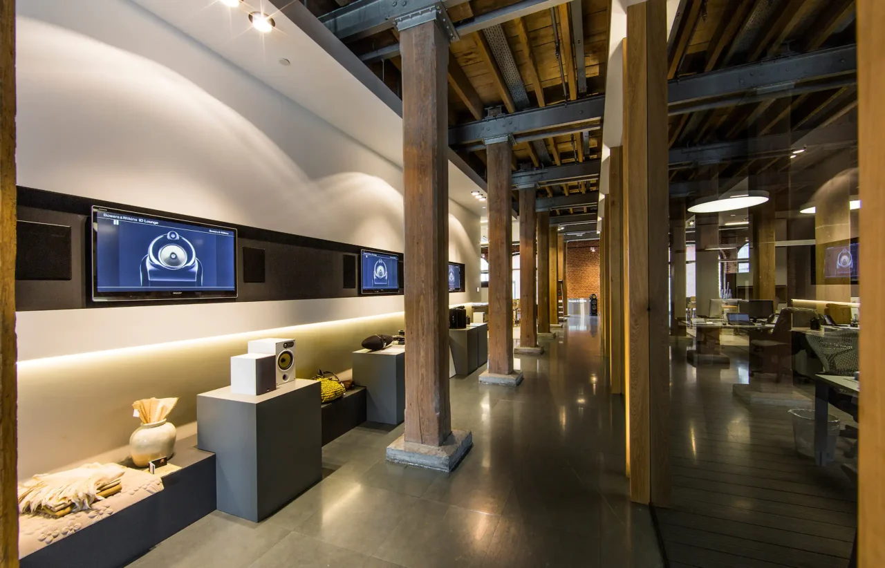 Interieur van de Bowers & Wilkins showroom door ERIC KANT met een gang voorzien van multimedia-displays, houten kolommen en een doorkijk naar de kantoorruimte, plus een luxe toiletruimte met bronzen deuren en donkere stucwanden.