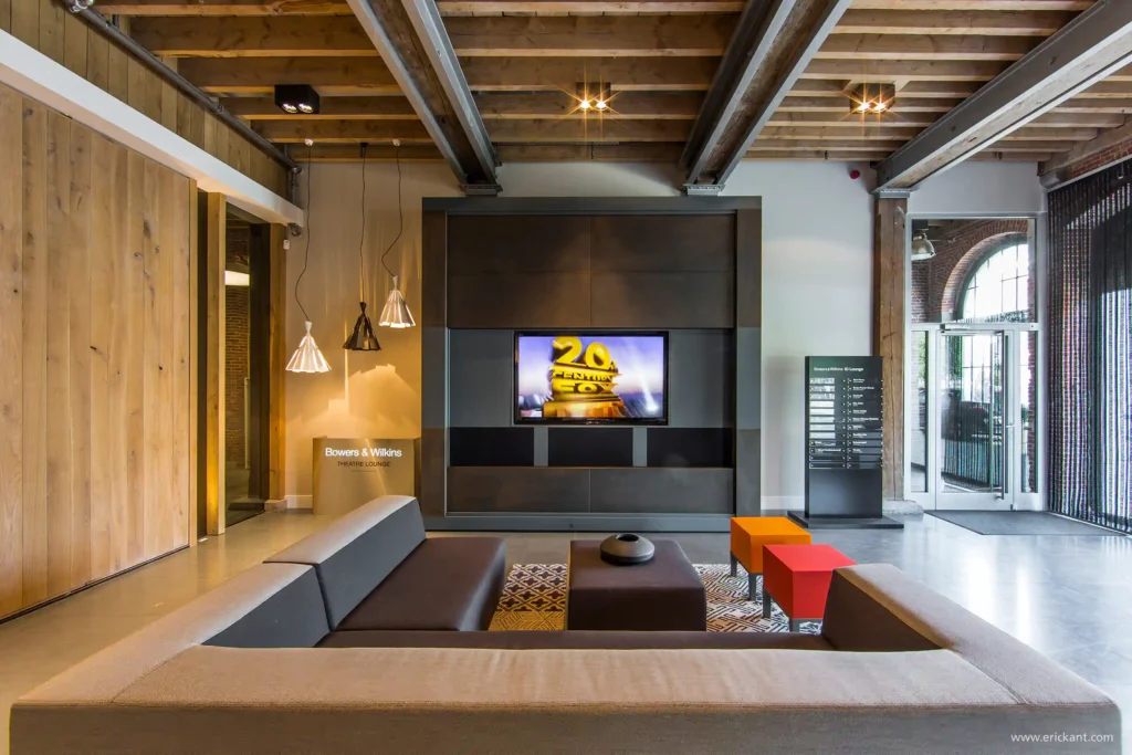 Interieur van de Bowers & Wilkins Theatre Lounge door ERIC KANT met een groot geïntegreerd bioscoopscherm, een moderne loungebank, kleurrijke poefs en een robuust balkenplafond.