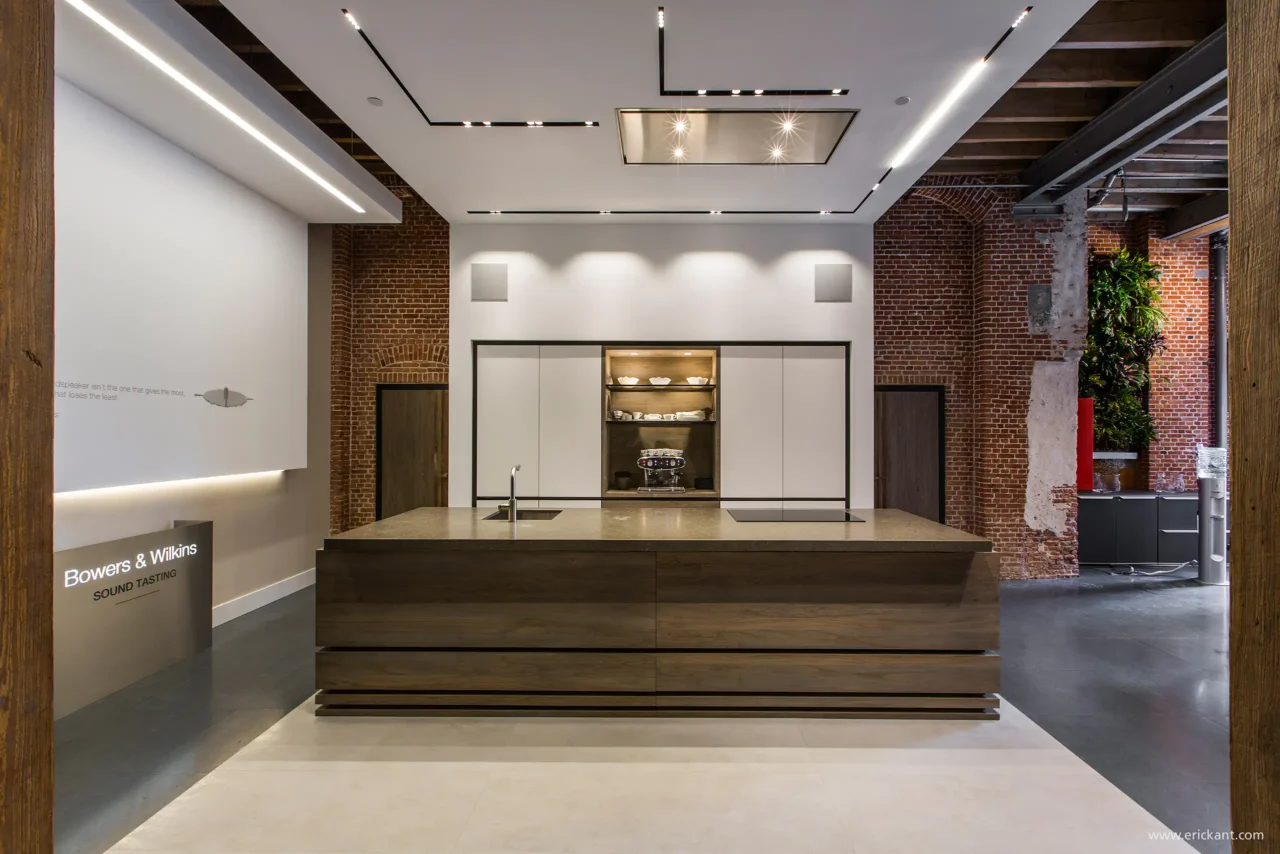 Een luxe keuken in de Bowers & Wilkins showroom door ERIC KANT met een groot houten kookeiland, een witte kastenwand, geïntegreerde audio en een modern lichtplan tegen een achtergrond van authentieke bakstenen muren.