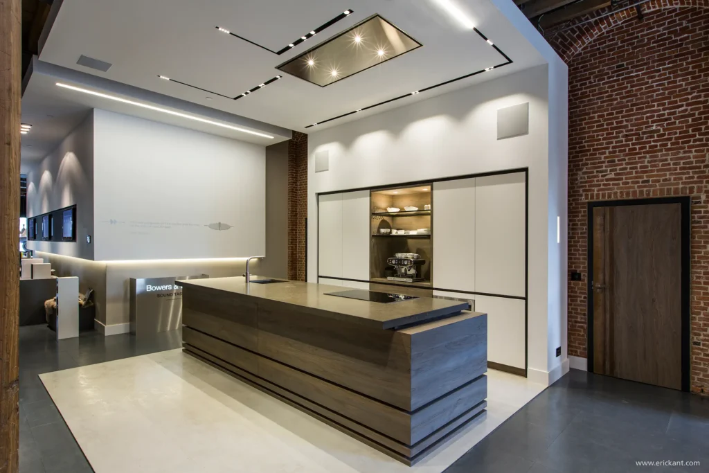 Perspectief van een luxe showroomkeuken door ERIC KANT met een massief houten kookeiland, geïntegreerde apparatuur, witte kastenwanden en een modern lichtplan in een industrieel pand.