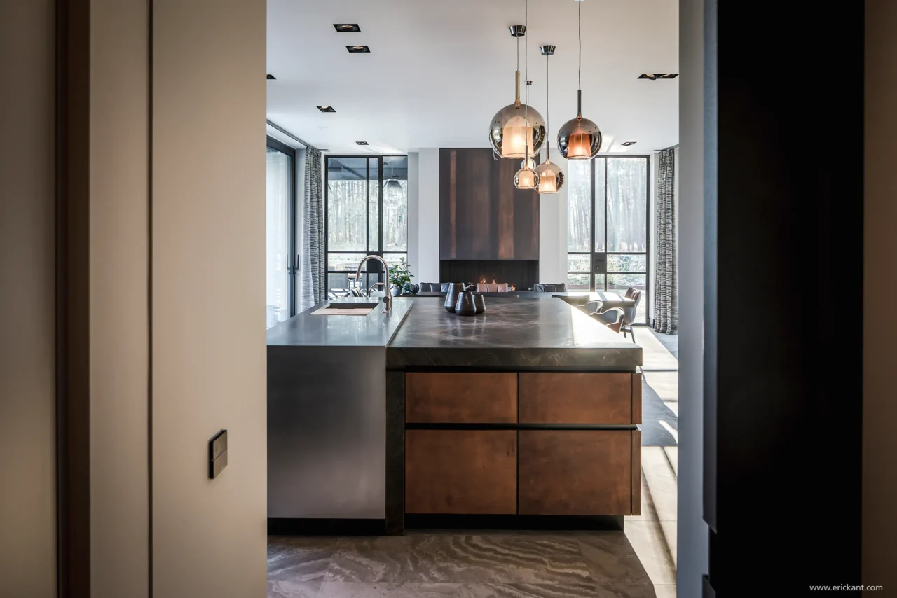 High-end keuken van de Modern Manor met een robuust kookeiland voorzien van bronzen fronten, een natuurstenen blad, mondgeblazen hanglampen en een doorkijkhaard door ERIC KANT.