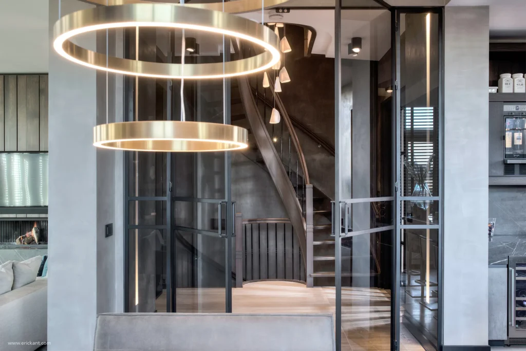 Interieur van de hal in de Mansion on the Waterfront door ERIC KANT met een imposante houten trap, stalen taatsdeuren, een moderne kroonluchter en zicht op de luxe living.Interieur van de hal in de Mansion on the Waterfront door ERIC KANT met een imposante houten trap, stalen taatsdeuren, een moderne kroonluchter en zicht op de luxe living.