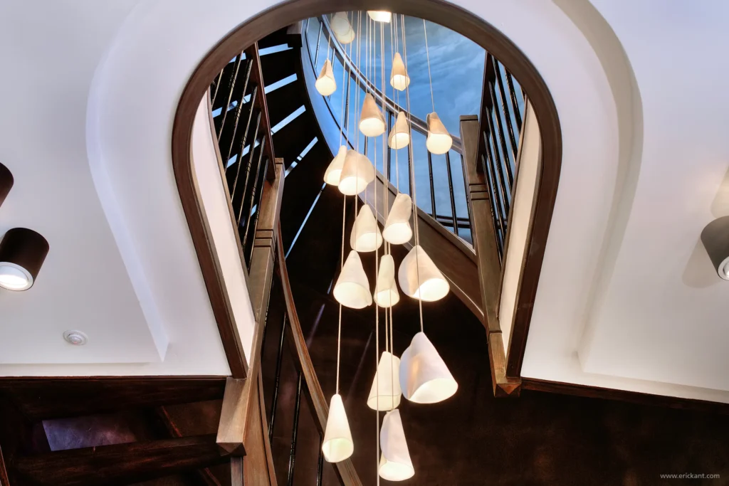 Close-up van een indrukwekkende cascade kroonluchter in het trappenhuis van de Mansion on the Waterfront door ERIC KANT, met witte organische kappen en zicht op de donkere houten trap en gestuukte plafonds.