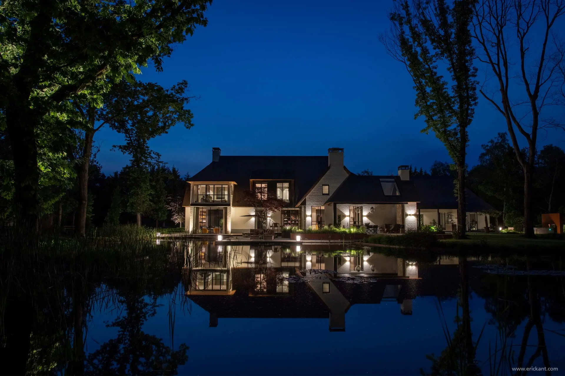 Nachtopname van de Modern Manor met een rieten dak en witte gevels, perfect gespiegeld in het water van een natuurlijke vijver door ERIC KANT.