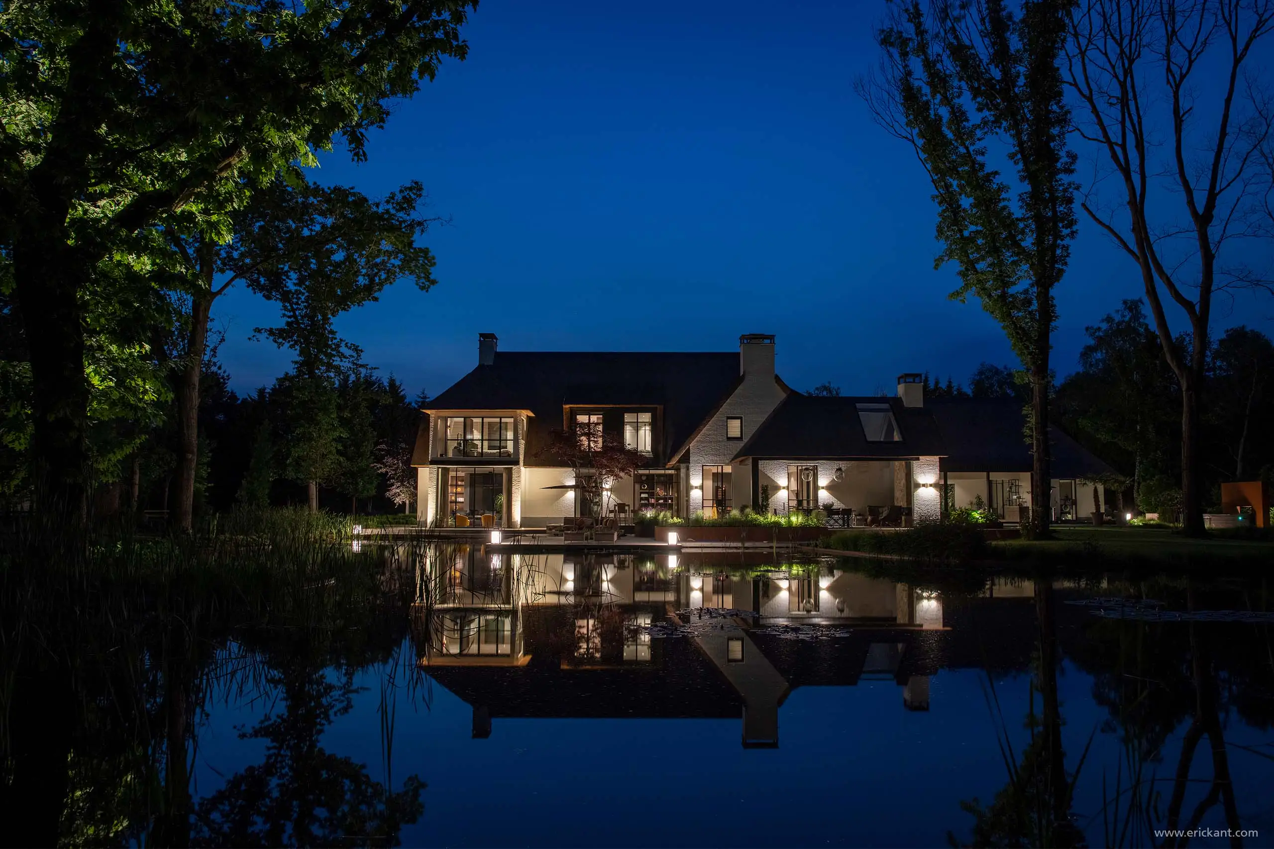 Nachtopname van de Modern Manor met een rieten dak en witte gevels, perfect gespiegeld in het water van een natuurlijke vijver door ERIC KANT.