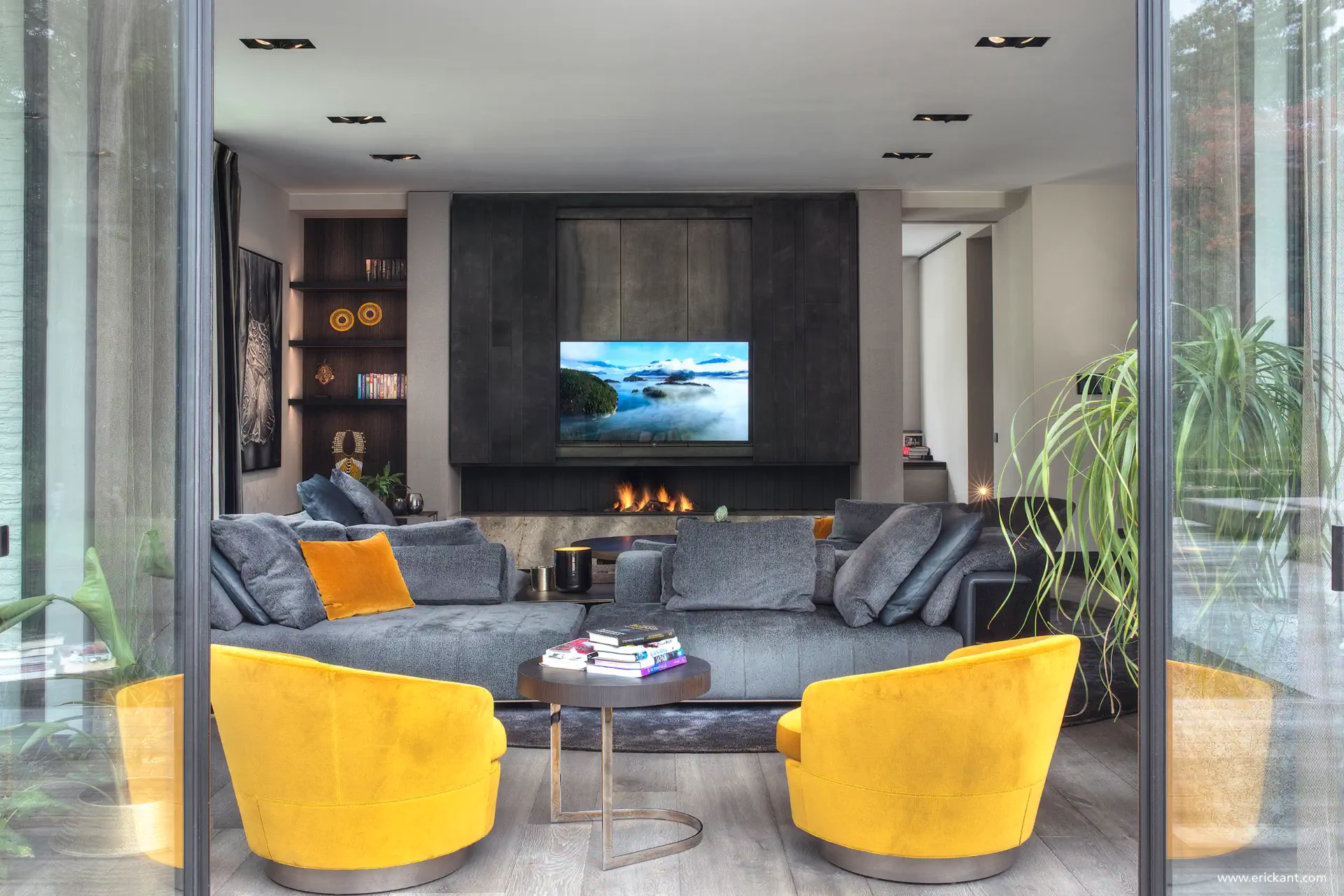 High-end interieur van de living in de Modern Manor met een grote grijze hoekbank, okergele fauteuils, een doorkijkhaard en een ingebouwde tv-wand door ERIC KANT.