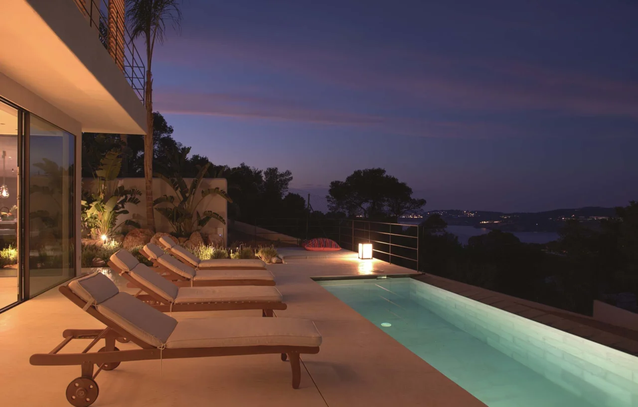 High-end zwembad bij nacht met houten ligbedden, sfeerverlichting en panoramisch uitzicht op de baai in project Cali Moli Ibiza door ERIC KANT.