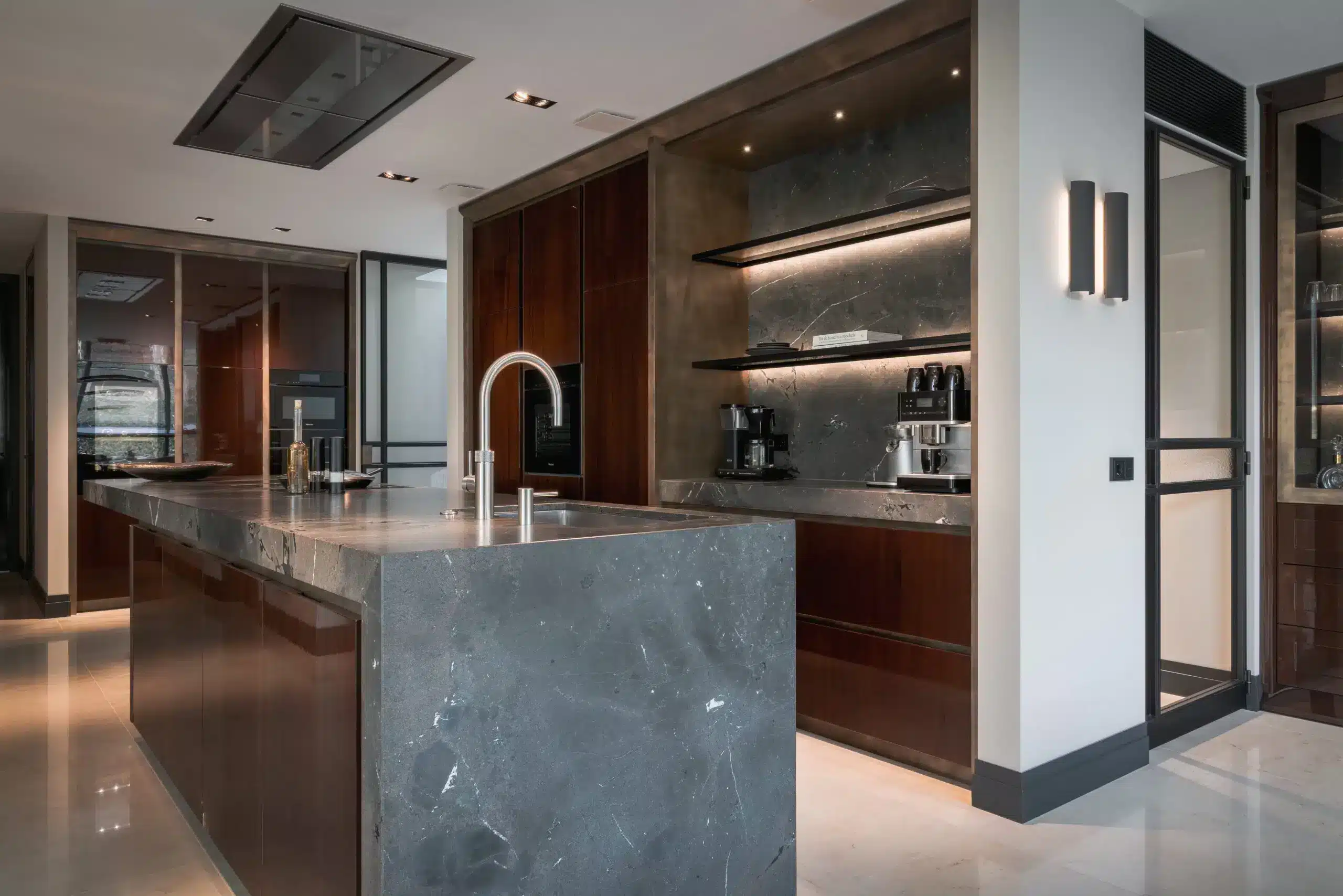 High-end keukeninterieur van de Modern Classic Villa met een massief natuurstenen kookeiland, hoogglans mahoniehouten kasten, een verlichte koffienis en een rvs designkraan door ERIC KANT.