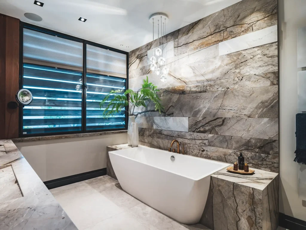 Exclusieve badkamer ontworpen door Eric Kant, met een indrukwekkende natuurstenen wand, een modern wit vrijstaand bad, bronzen kranen en een stijlvolle hanglamp.