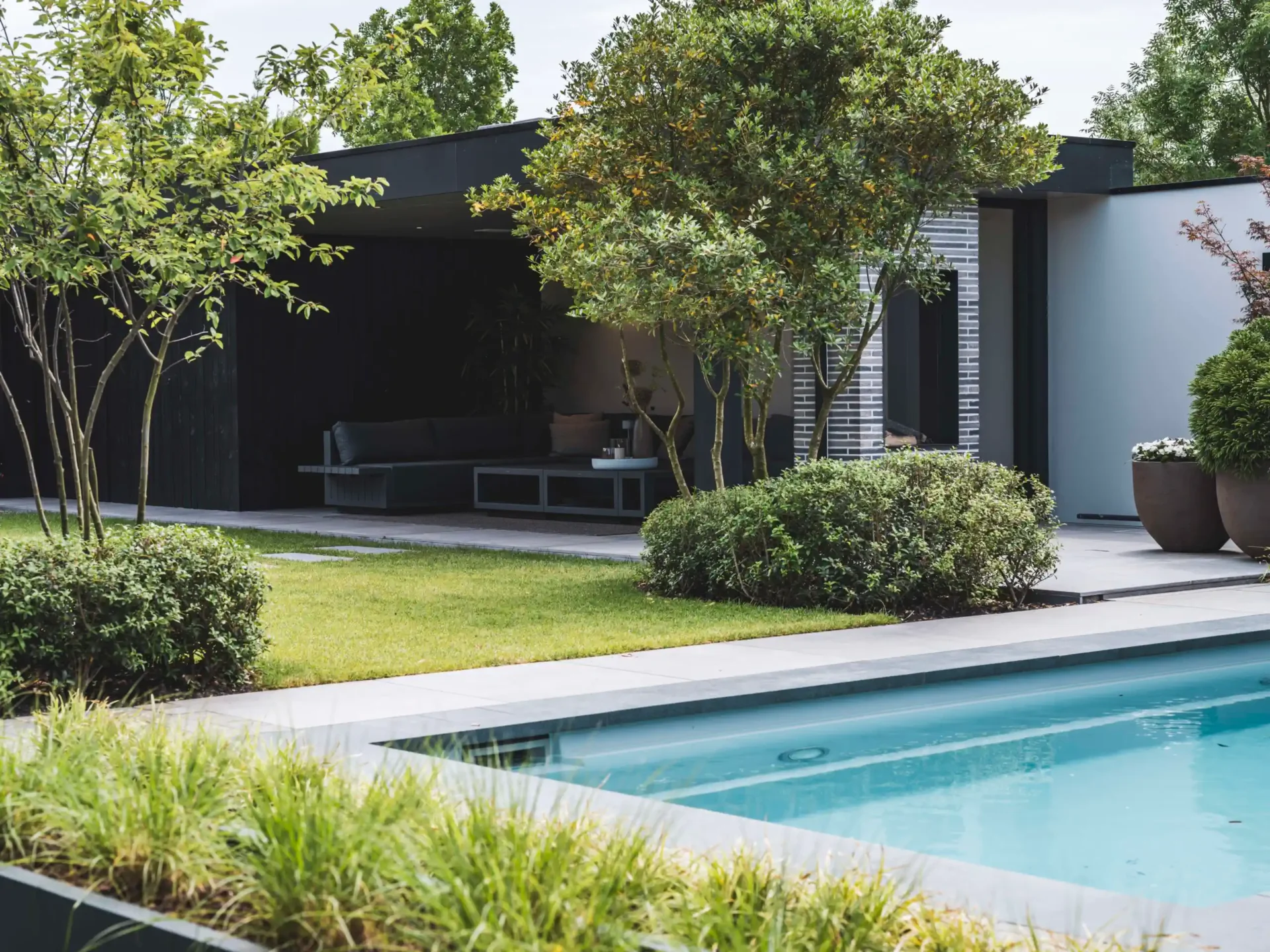 Modern tuinontwerp door Eric Kant, met een strak zwembad op de voorgrond, een groen gazon en een luxe zwart houten buitenverblijf met comfortabele loungehoek.