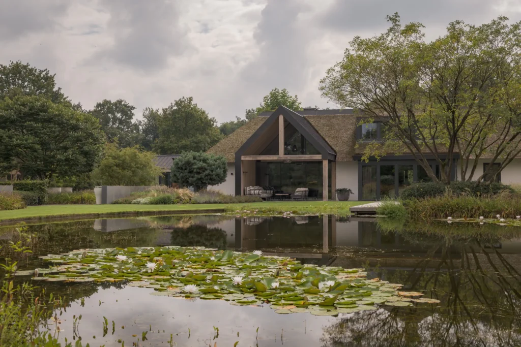 Exterieur van een exclusieve rietgedekte villa in Erp door ERIC KANT, met een grote glazen gevel, eikenhouten spanten, een strak gazon en een natuurlijke vijver met waterlelies.