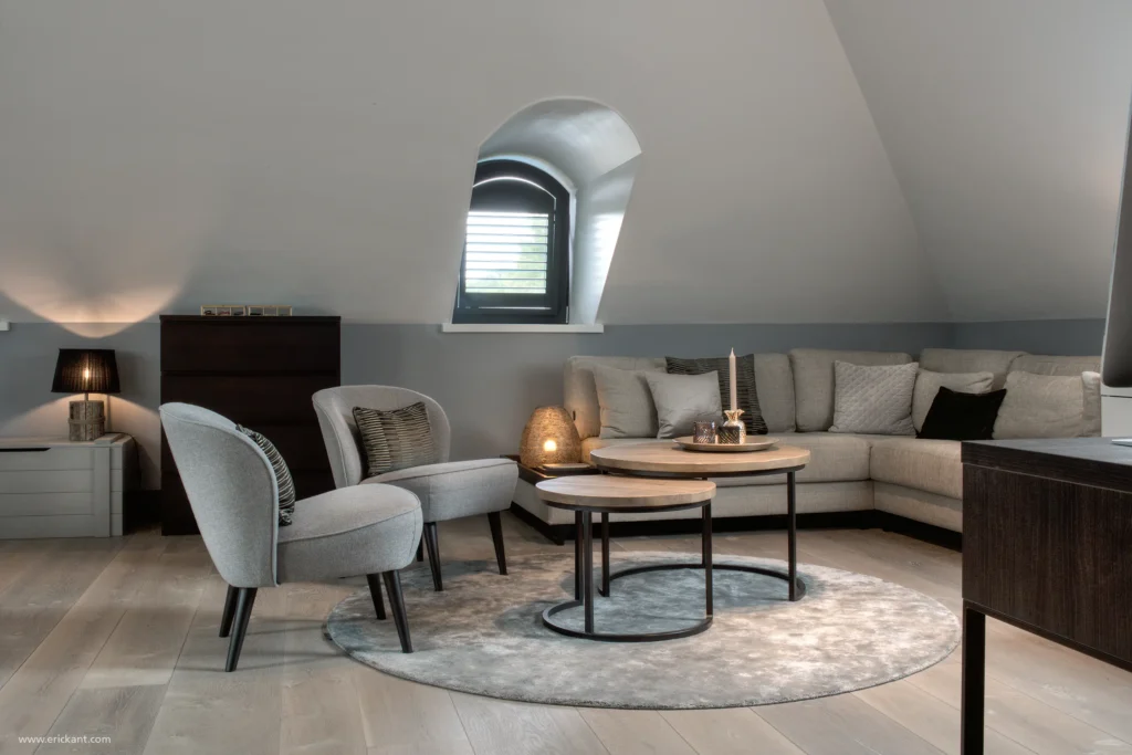 Interieur van een luxe gastenverblijf in de Manor Villa door ERIC KANT, gesitueerd onder de kap met een rond dakkapelraam, een comfortabele hoekbank, stijlvolle fauteuils en een houten vloer.