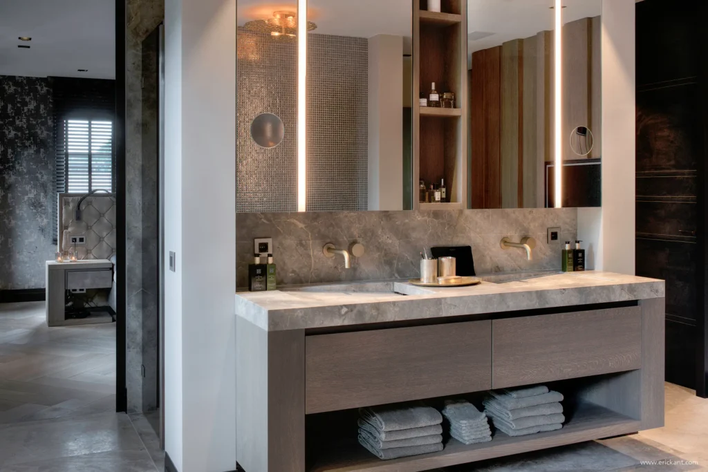 High-end badkamerinterieur in de Manor Villa door ERIC KANT met een dubbel wastafelmeubel van eikenhout, een massief natuurstenen blad met geïntegreerde kranen, verlichte spiegels en zicht op de aangrenzende master bedroom.