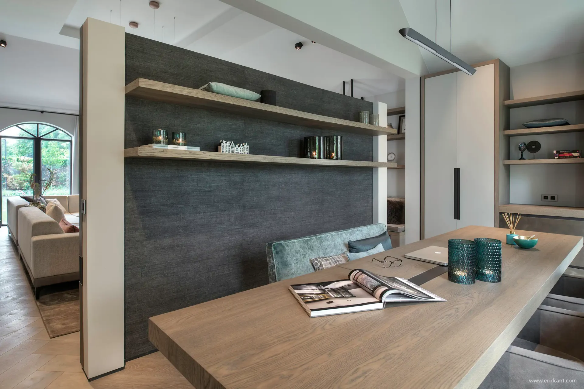 High-end thuiskantoor in de Manor Villa door ERIC KANT met een robuust eikenhouten bureau, maatwerk wandplanken op een textielwand, een fluwelen bank en zicht op de aangrenzende living.