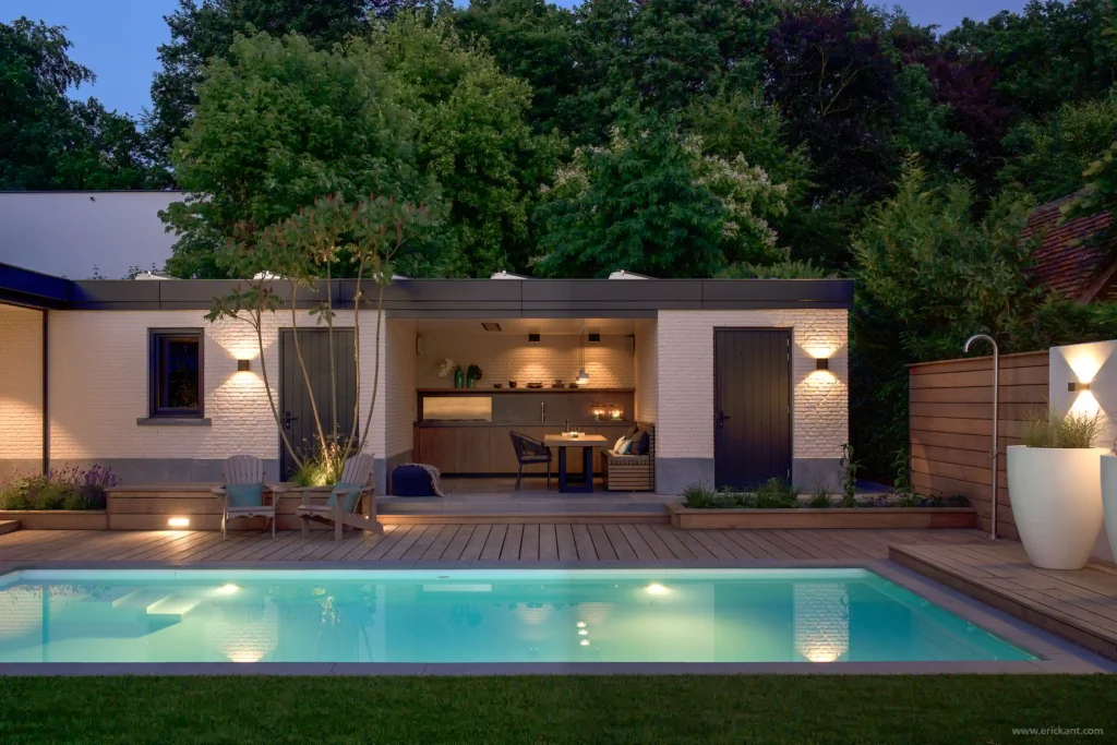 High-end zwembad en pool house van de Manor Villa door ERIC KANT met een houten deck, een luxe buitenkeuken, sfeervolle gevelverlichting en een strakke tuinarchitectuur bij schemering.