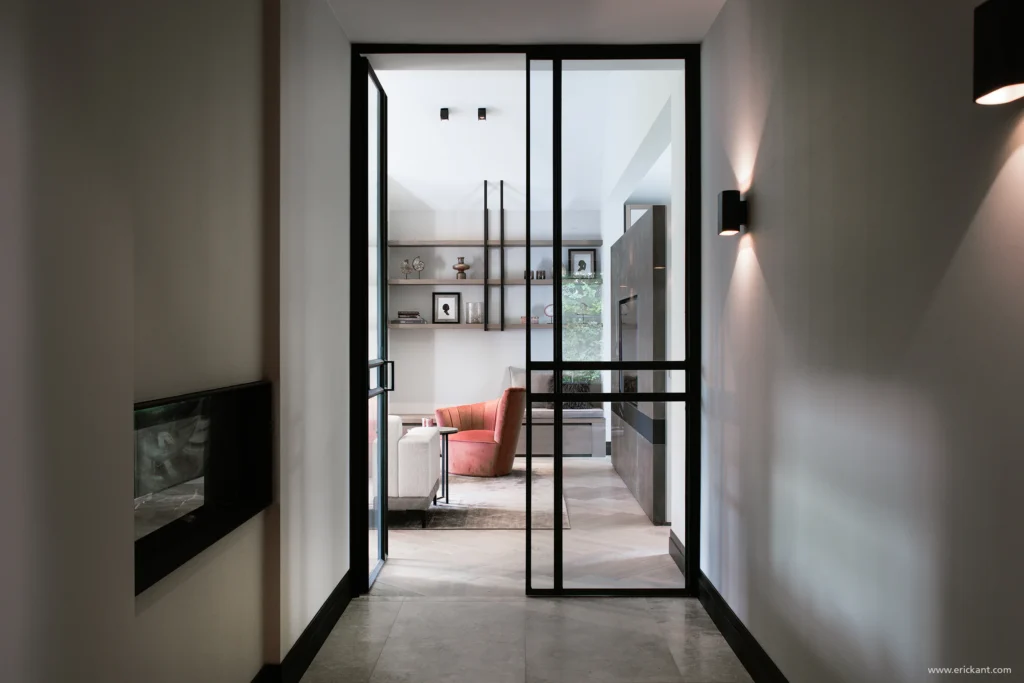 Zicht vanuit de hal van de Manor Villa door ERIC KANT door zwarte stalen deuren naar de verlichte living, met zicht op een visgraatvloer, een oudroze fauteuil en maatwerk wandplanken.