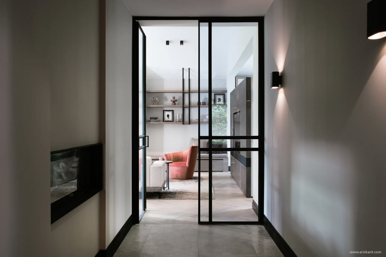 Zicht vanuit de hal van de Manor Villa door ERIC KANT door zwarte stalen deuren naar de verlichte living, met zicht op een visgraatvloer, een oudroze fauteuil en maatwerk wandplanken.