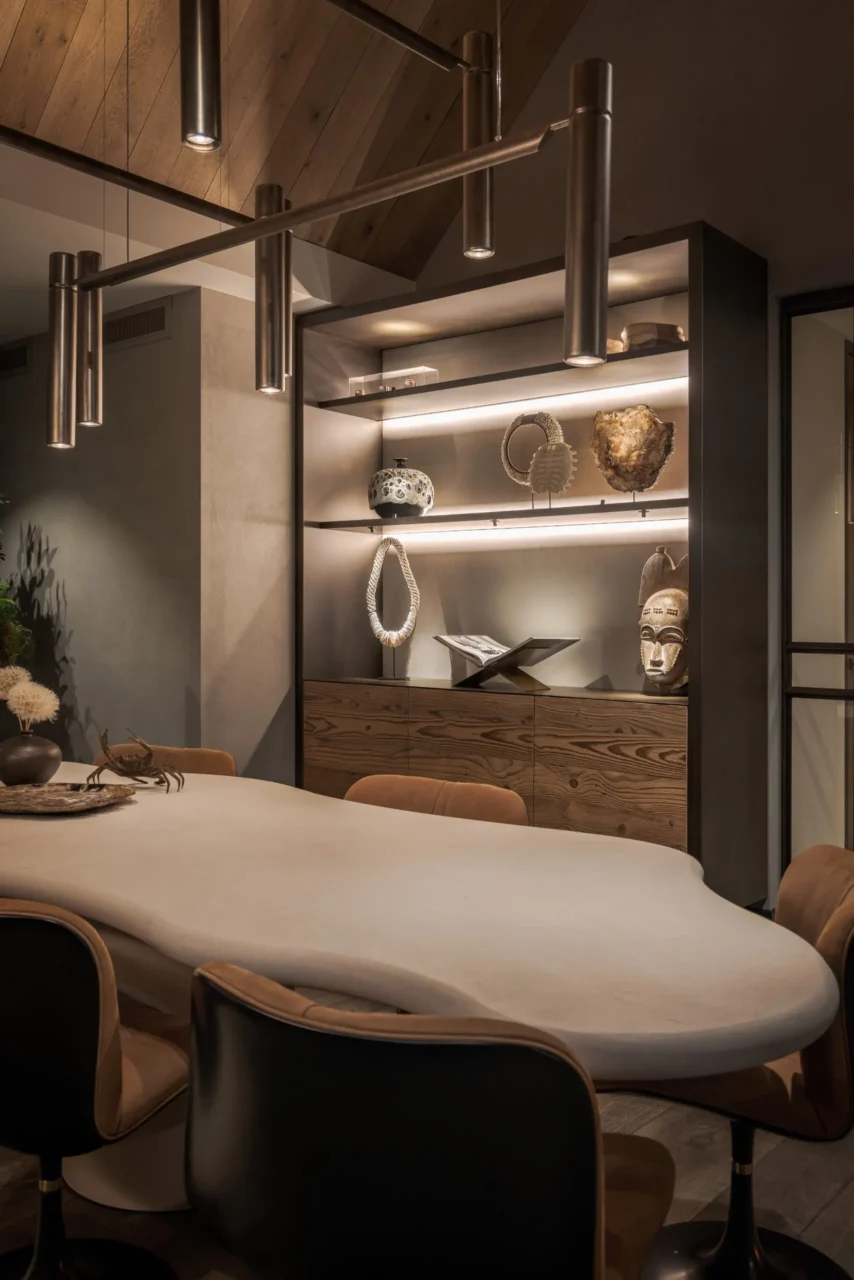 Detail van een luxe eetkamer ontworpen door Eric Kant, met een maatwerk dressoir van robuust hout, verlichte nissen met etnische kunstobjecten, een organisch gevormde eettafel en moderne bronzen hangverlichting.