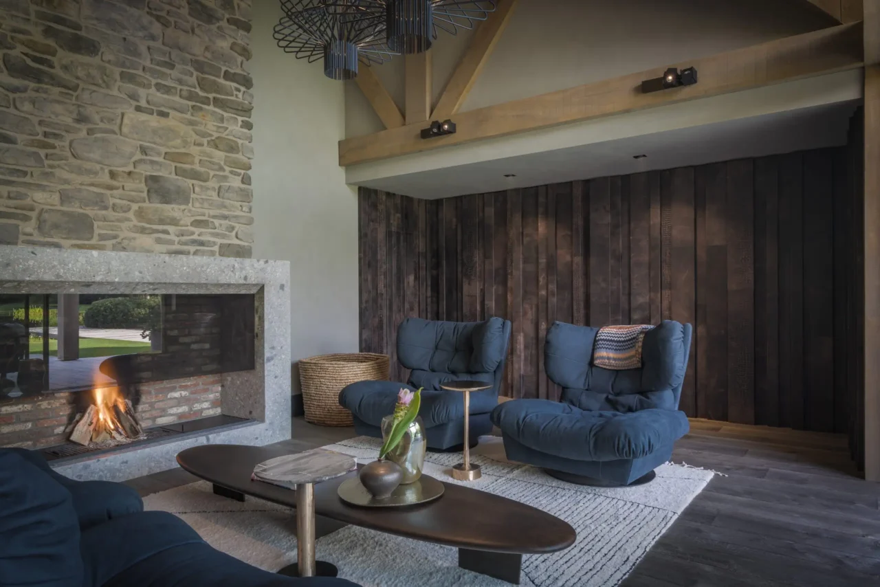 Exclusieve woonkamer in African Lodge stijl ontworpen door Eric Kant, met Arflex Strips fauteuils, een robuuste natuurstenen haardwand, houten balken en een sfeervol vuur.