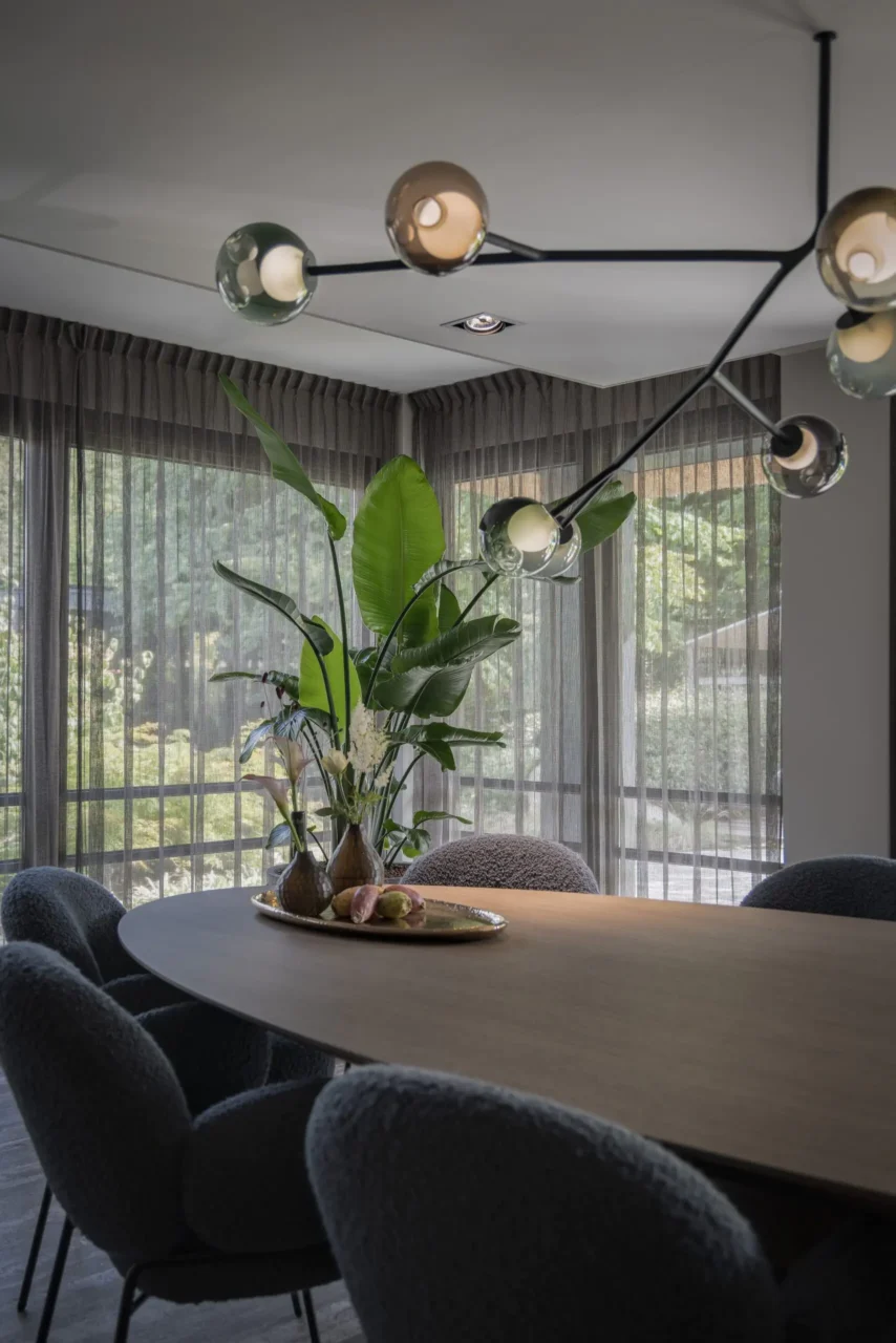 Interieurontwerp van een luxe eetkamer door Eric Kant, met een ovale houten tafel, grijze bouclé stoelen, een moderne zwarte hanglamp met glazen bollen en grote kamerplanten bij het raam.Interieurontwerp van een luxe eetkamer door Eric Kant, met een ovale houten tafel, grijze bouclé stoelen, een moderne zwarte hanglamp met glazen bollen en grote kamerplanten bij het raam.