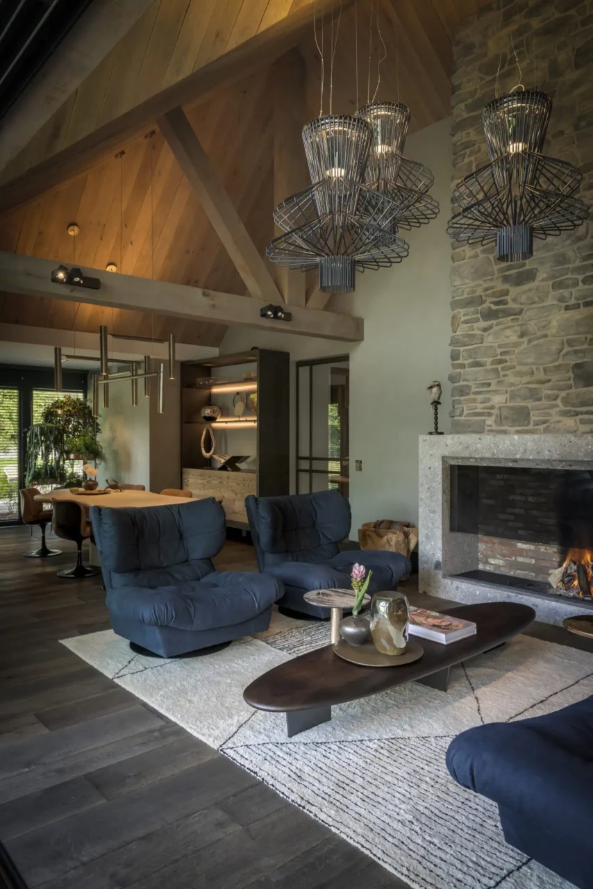 Exclusieve woonkamer ontworpen door Eric Kant, met Baxter Milano fauteuils, een robuuste natuurstenen haardwand en een hoog houten plafond.