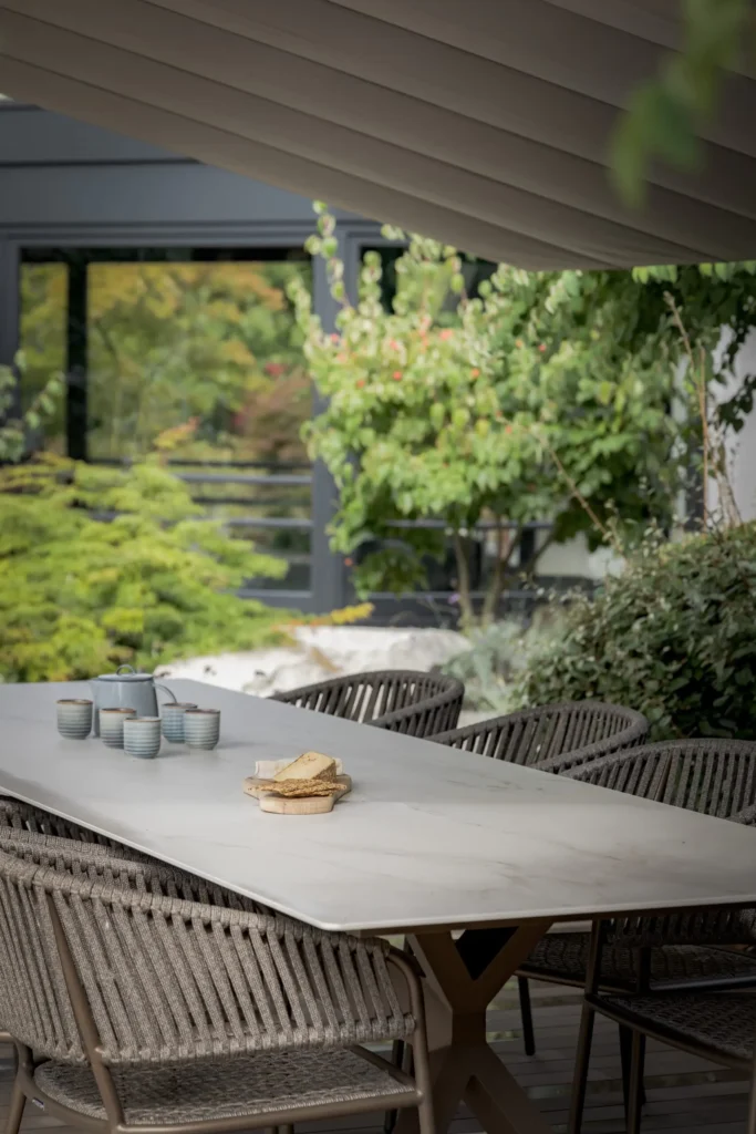 Detail van een luxe buitenterras ontworpen door Eric Kant, met een grote keramische eettafel, gevlochten design tuinstoelen en sfeervol servies, omgeven door groen.